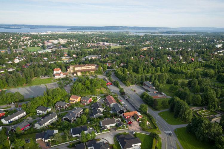 Luftbilde av et boligområde med grønne trær, veier og hus i nærheten av en innsjø.