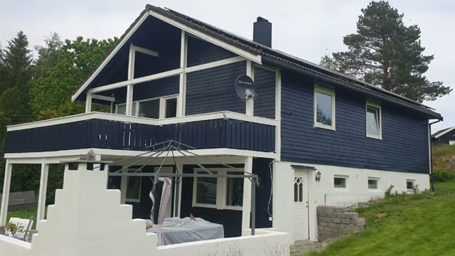 Blått hus med hvite detaljer, terrasse og hage, omgitt av trær og grønt landskap.