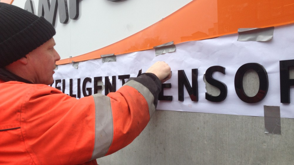 En mann i oransje arbeidsjakke fester et skilt med teksten "INTELLIGENT SENSOR" på en vegg.