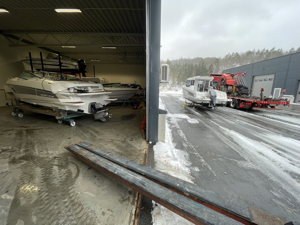 Båter i en verkstedhall med snø på bakken og en lastebil som laster en båt utenfor.