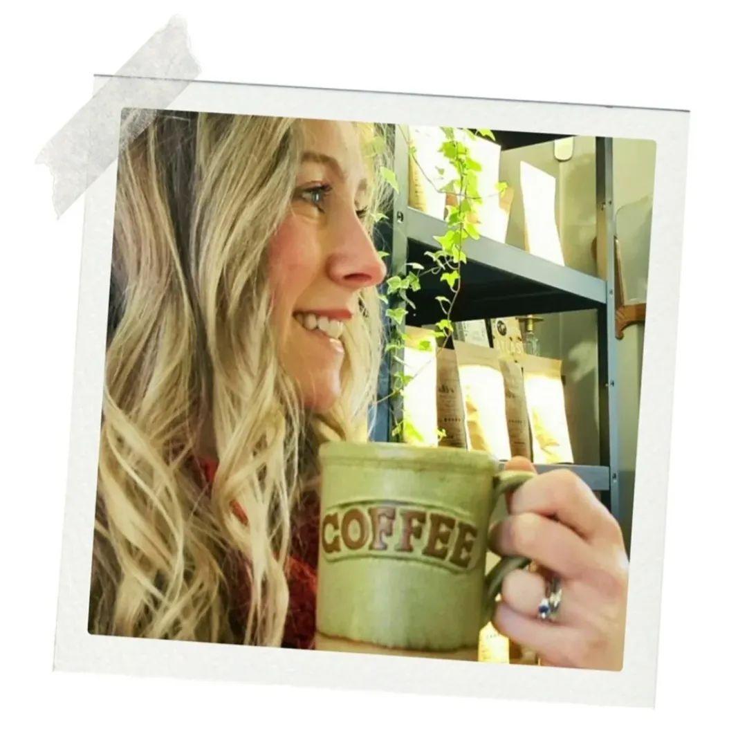 Kvinne med langt hår holder en kaffekopp i en koselig kafé med planter i bakgrunnen.