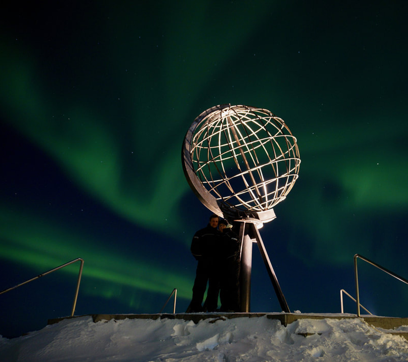 En person står ved et globusmonument under nordlys på en snødekt plattform.