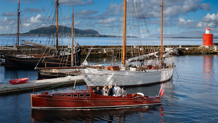 Båter i havnen med en rød seilbåt og en stor hvit seilbåt, omgitt av fjell og blå himmel.