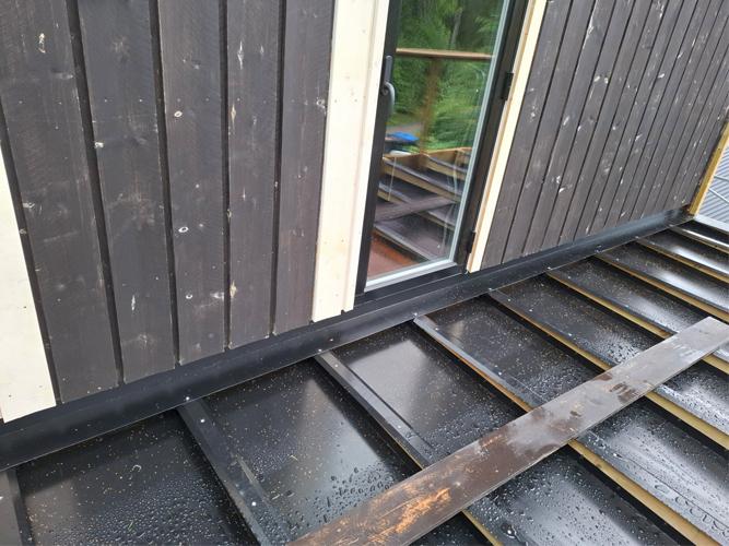 Tak med mørke planker og vindu, med en treplanke liggende på det våte underlaget.