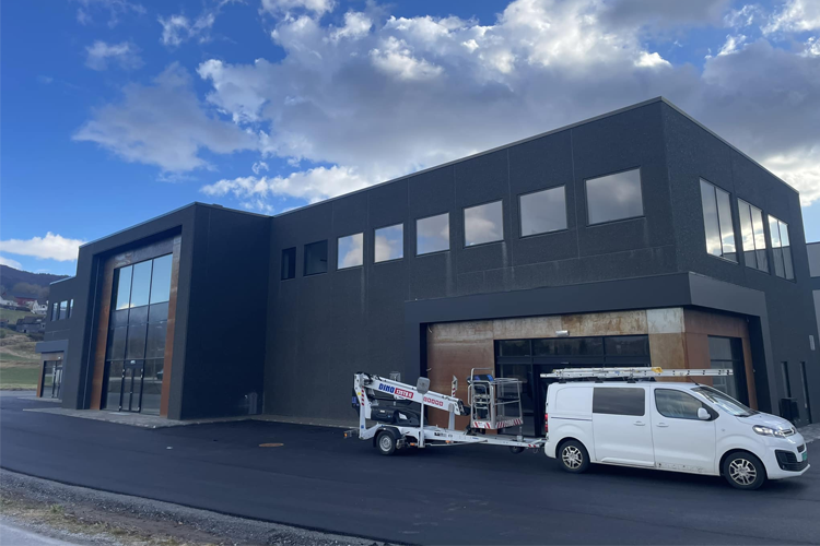 Moderne bygning med store vinduer og en hvit varebil parkert foran, med en tilhenger med utstyr.