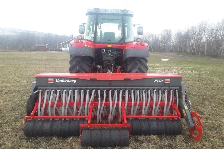 Rød traktor med såmaskin på en åpen eng, omringet av trær og fjell i bakgrunnen.