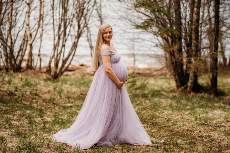 Gravid kvinne i en lang, lavendelfarget kjole står i en skog med trær og vann i bakgrunnen.