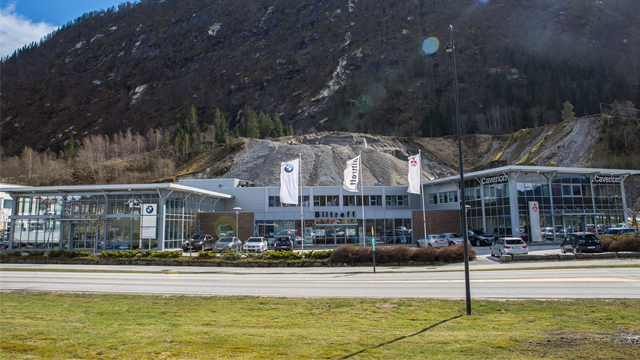 Bilforhandler med moderne bygning og flagg, beliggende ved veien med fjell i bakgrunnen.