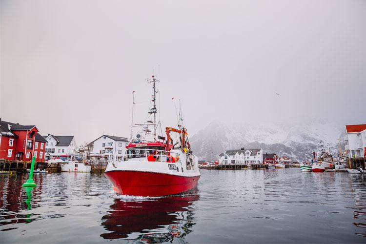 En rød fiskebåt seiler inn i havnen med hvite hus og fjell i bakgrunnen. Vannflaten speiler omgivelsene.