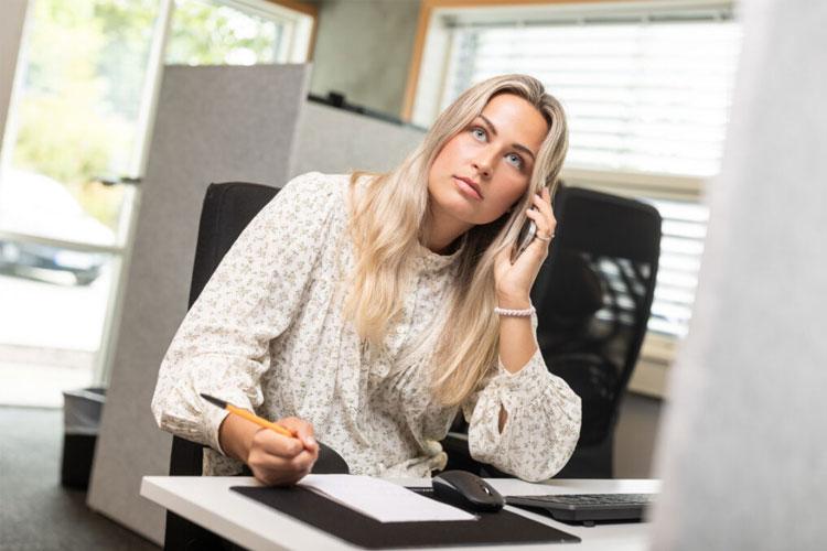 Kvinne med langt hår som snakker i telefonen mens hun skriver notater på et kontor.