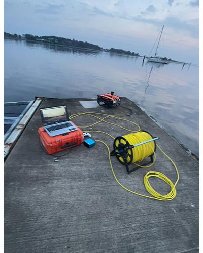 Utrustning på en brygge ved vannet, inkludert en oransje boks, en gul kabelrull og en liten båt i bakgrunnen.