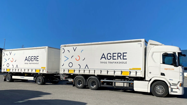 En lastebil med tilhenger, merket med "AGERE", står parkert under en klar blå himmel.