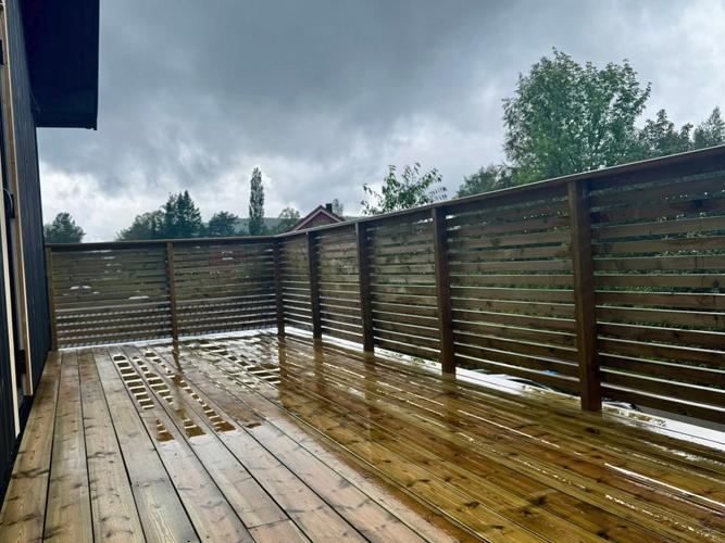 En våt terrasse med treplanker og en gjerde av tre, under en mørk sky med trær i bakgrunnen.