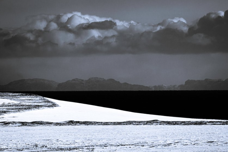 Et svart-hvitt bilde av en snødekt landskap med mørke skyer og fjell i bakgrunnen.