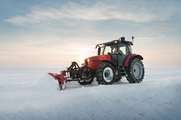 Rød traktor som pløyer snø på en vinterlig landskap med soloppgang i bakgrunnen.
