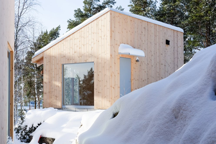 Moderne hytte med trepanel og store vinduer, omringet av snø og trær i en vinterlig setting.