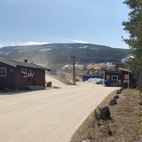 Utsikt over en vei med røde bygninger og fjell i bakgrunnen, omgivelsene er solfylte og støvete.