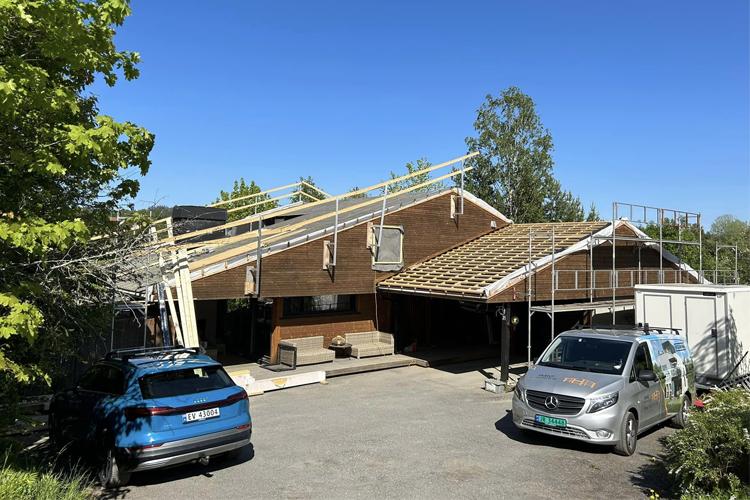 Hus med takarbeid, stillas og biler parkert foran, omkranset av trær under klar blå himmel.