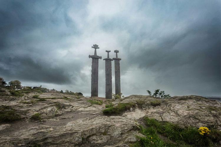 Tre store sverd står på en steinete bakgrunn under en mørk, skyet himmel.