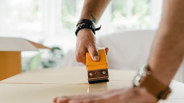 En person bruker en gul tape-dispenser for å feste tape på en pappeske.
