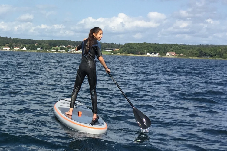 En jente står på et paddleboard i vannet, iført våtdrakt, med en padleåre i hånden.