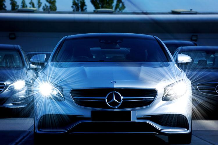 En sølvfarget Mercedes-bil med lysene på, parkert blant andre biler i en moderne setting.
