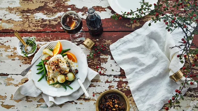 En tallerken med fisk, grønnsaker og sitrusfrukter, servert med et glass brus og dekorative elementer på bordet.