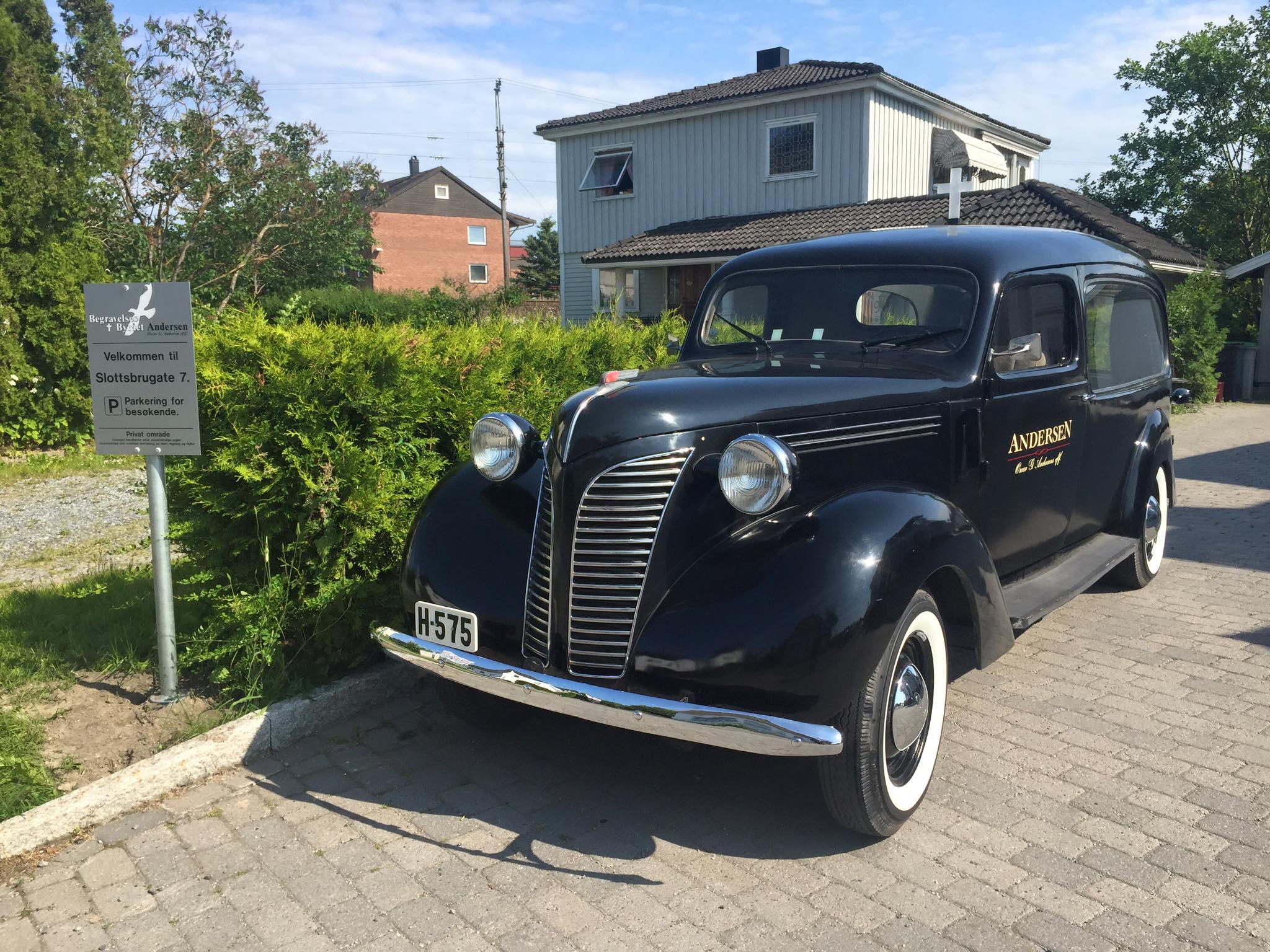 En svart veteranbil fra 1930-tallet parkert foran et hus med grønt buskas og skilt i bakgrunnen.