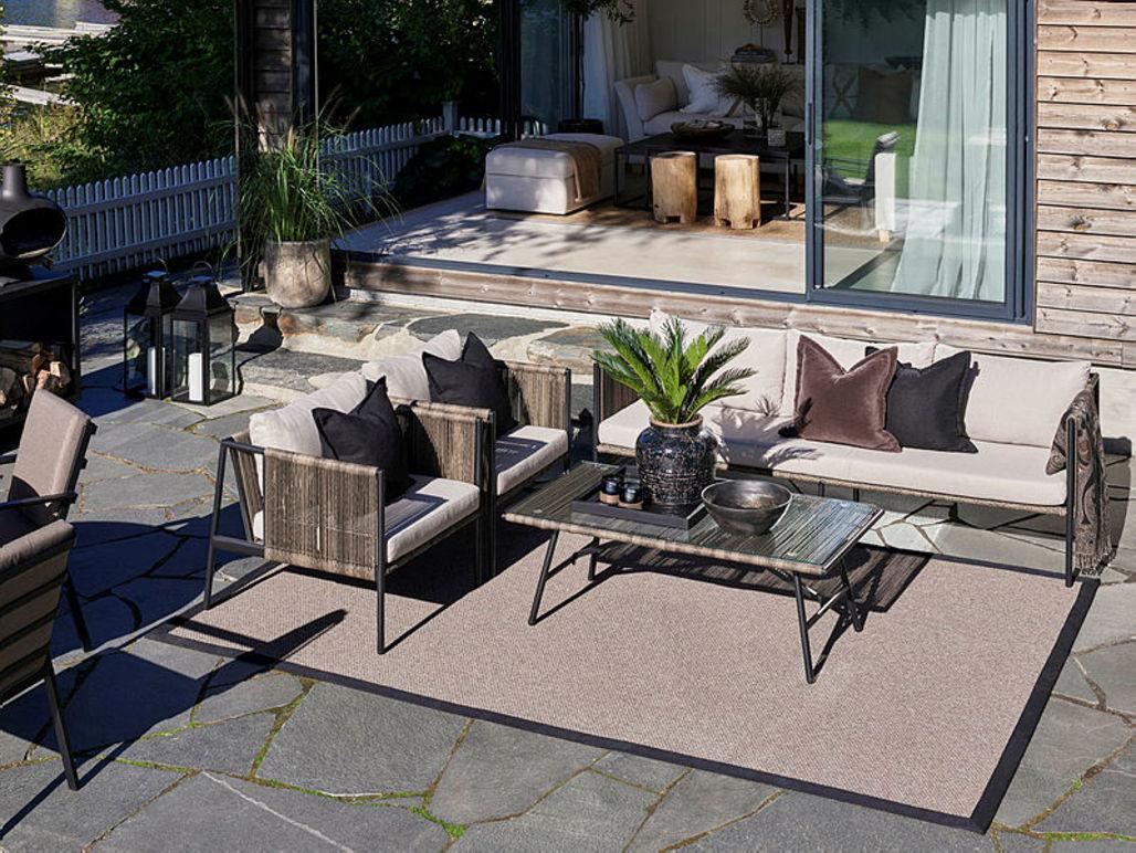 Uteplass med moderne møbler, inkludert sofa, stoler og bord, omgitt av planter og en terrasse.