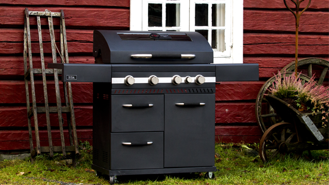 En moderne gassgrill med fire brennere, plassert foran en rød trevegg med vindu.