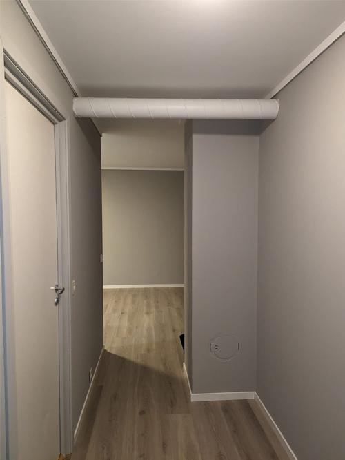 En lys gang med grå vegger og lyst gulv, som fører til et rom i bakgrunnen.