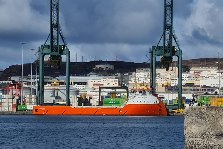 Fartøy i havn med kraner og containere i bakgrunnen, under en skyet himmel.