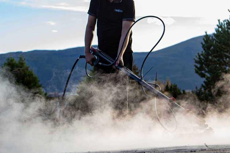 En person bruker en dampmaskin på en vei, med damp som stiger opp i luften og fjell i bakgrunnen.