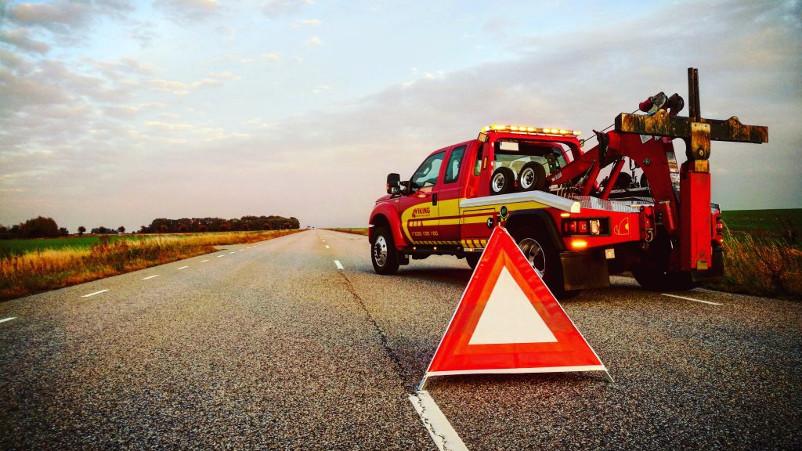 En rød bergingsbil står på veien med et varseltrekanter foran, omringet av åkerlandskap.