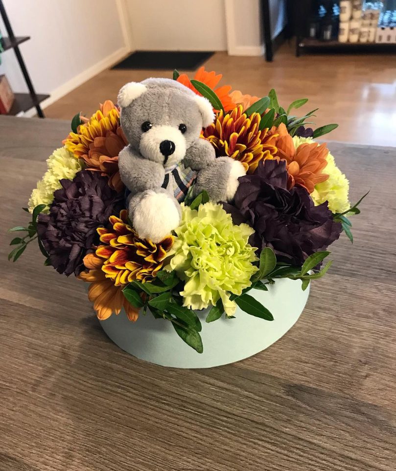 En blomsteroppsats med en grå bamse i midten, omringet av fargerike blomster som chrysantemum og nelliker.