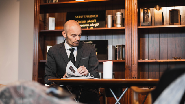 En mann i dress sitter ved et bord og leser en brosjyre i et rom med trevegger og dekorative gjenstander.