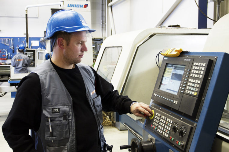 En mann i blå hjelm og grå vest betjener en CNC-maskin i en moderne fabrikk.