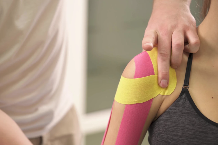 En person får kinesiotape på skulderen, med rosa og gul tape som støtter muskulaturen.