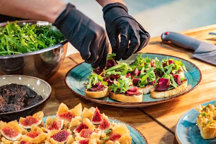 En person med hansker lager småretter med brød, salat og rødbeter på et bord med flere skåler med ingredienser.