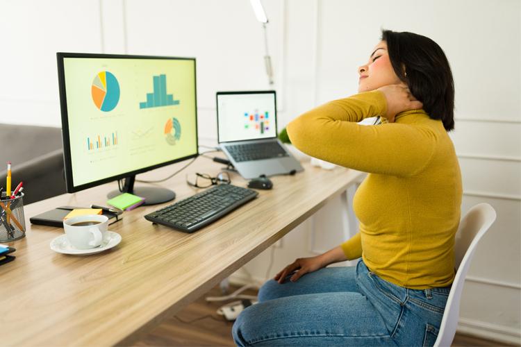 En kvinne sitter ved et skrivebord med datamaskiner, hun holder seg til nakken og ser ubehagelig ut.