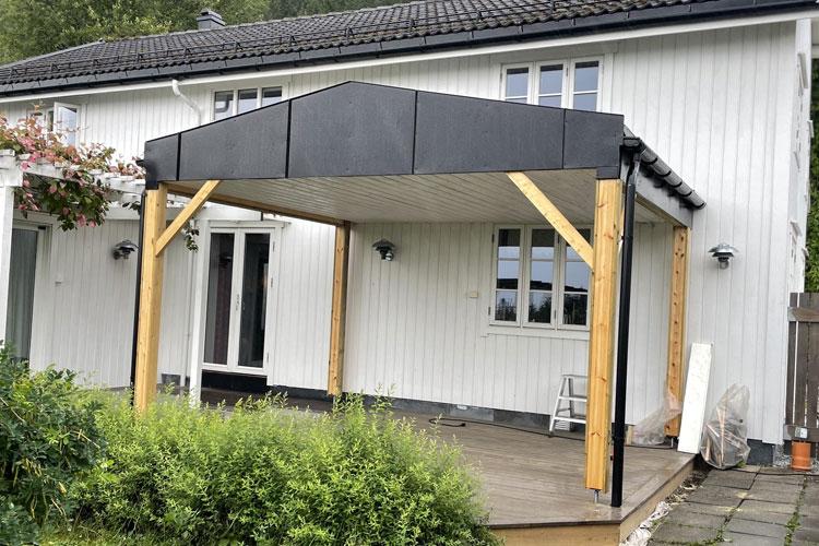 Uteplass med tak over terrasse, bygget i tre, ved en hvit husvegg med vinduer.