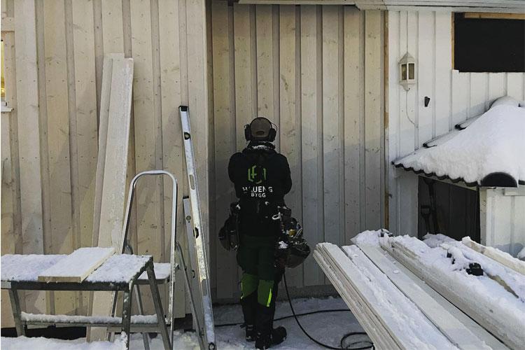 En håndverker står foran en vegg av tre, iført verneutstyr, med snø og byggeplanker rundt seg.