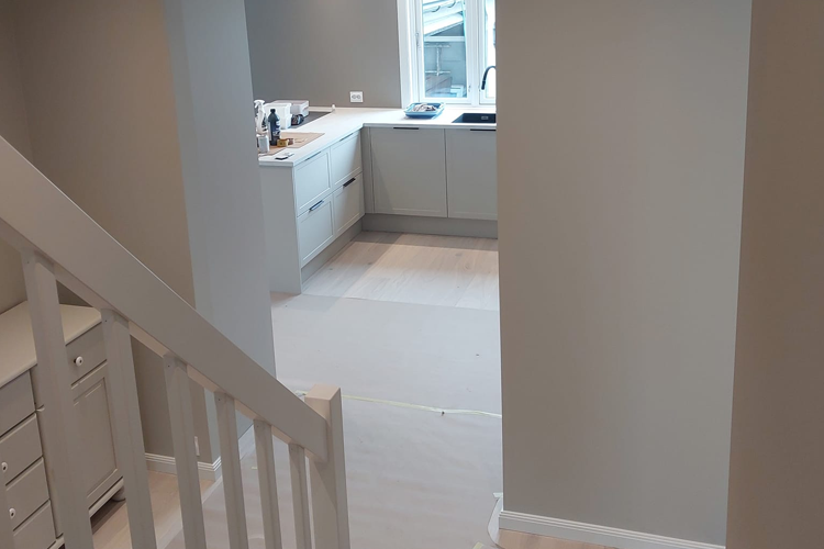 Moderne kjøkken med grå vegger, hvite skap og trappen i forgrunnen. Lysinnslipp fra vinduet.