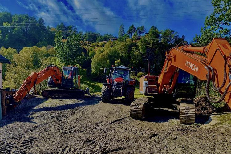 Tre gravemaskiner og en traktor står på en grusvei omringet av grønn natur og trær under en blå himmel.
