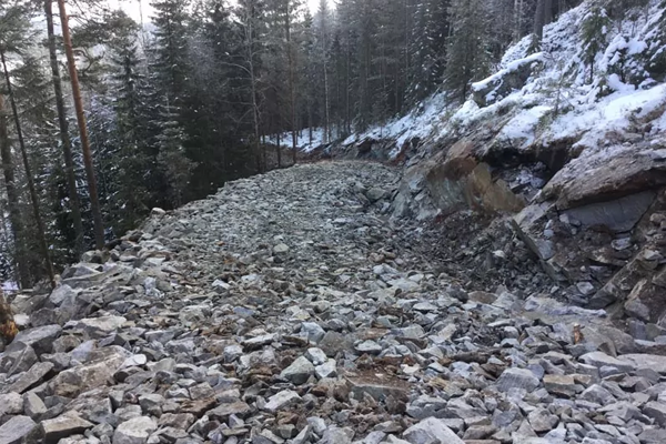 En steinete vei i skogen, omringet av trær og snø, med store steiner og grus på bakken.