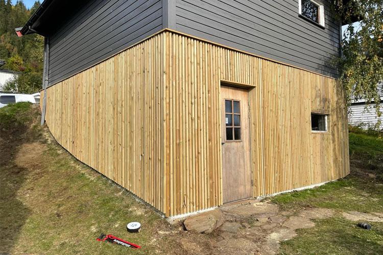 Hjørnet av en bygning med trepanel, dør og vindu, omgitt av grønt gress og verktøy på bakken.
