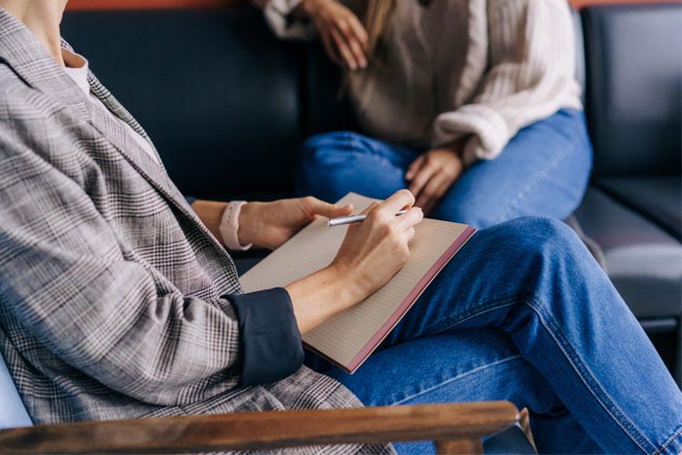 En person skriver i en notatbok mens en annen sitter i bakgrunnen på en sofa.