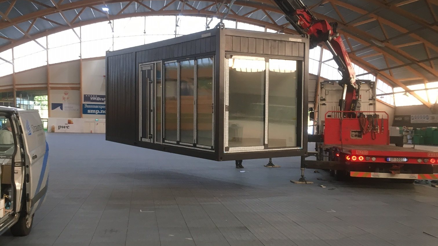 En kran løfter en container med store vinduer inne i en hall med takbjelker.