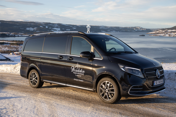 En svart Mercedes V-Klasse minivan parkert ved en snødekt vei med fjell og innsjø i bakgrunnen.