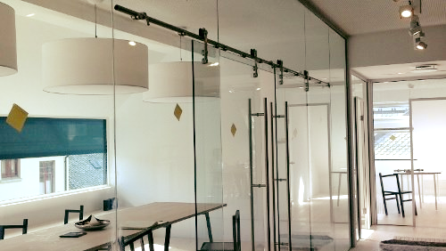 Moderne kontor med glassvegger, taklamper og en stor bordplass i et lyst rom.